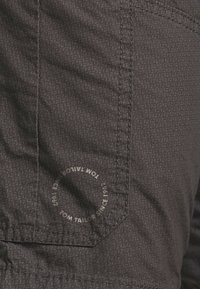Tejido oscuro y texturizado con un patrón sutil, que presenta un bolsillo cosido y un detalle de marca circular que dice "Tom Tailor Since 1961".