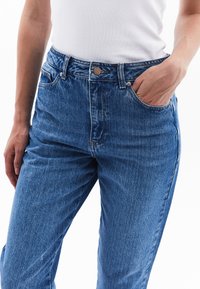 Korkeavyötäröiset siniset farkut, joissa on hieman kulunut pinta, napitus ja vetoketju, viisi taskua ja suora lahje.