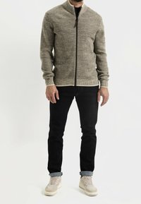 Giacca beige lavorata a maglia con zip, texture a coste, colletto alto e zip nera a contrasto. Abbinata a jeans scuri e sneakers chiare.