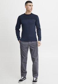 Marineblå strikkegenser med teksturert mønster, rund hals og kontrasterende horisontal stripe. Kombinert med grå bukser og svarte joggesko.