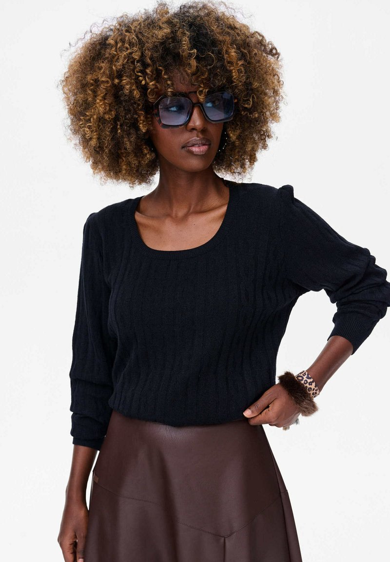 Czarny sweter z prążkowanej dzianiny, z szerokim dekoltem w serek i długimi rękawami, w zestawieniu z brązową skórzaną spódnicą. Dodatki obejmują bransoletkę w panterkę.