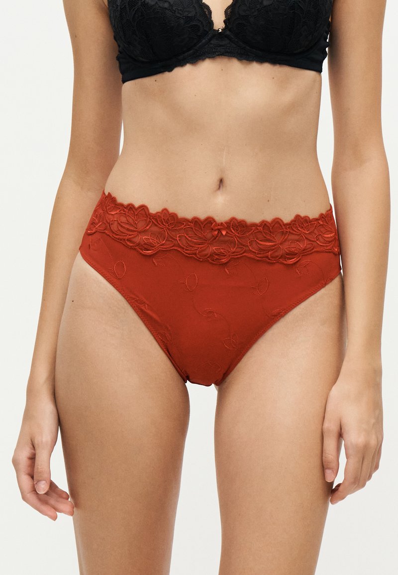 Femme portant un soutien-gorge en dentelle noire et un sous-vêtement rouge taille haute orné de dentelle, debout devant un fond uni.