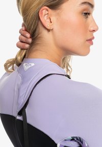 Violetti ja musta märkäpuku, jossa on korkeakauluksinen muotoilu, sileä pinta ja sydänlogo olkapäällä. Takana näkyvät hihnat.