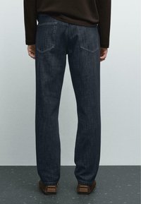 Dunkelblaue Jeans aus Denim mit geradem Bein. Verfügt über zwei Gesäßtaschen und eine dezente Textur. Getragen mit einem braunen Langarmoberteil.