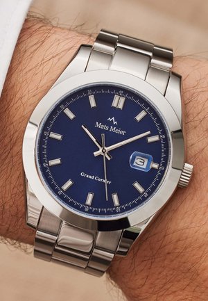 Montre en acier inoxydable argenté avec un cadran bleu, des index blancs, une fenêtre de date et un bracelet en métal brossé. Marque : Mats Meier, modèle : Grand Corner.