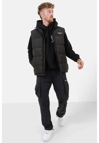 Zwarte gewatteerde vest met hoge kraag over een zwarte hoodie, gecombineerd met zwarte cargobroeken en witte sneakers. Model is aan het lopen.