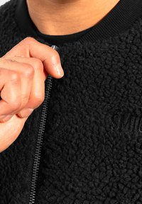 Mano agarrando el tirador de la cremallera de una chaqueta de forro polar texturizado negro con texto bordado parcialmente visible en el pecho.