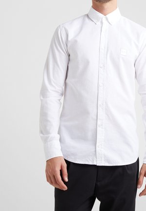 Hombre con camisa blanca ajustada de botones y mangas largas, y pantalones negros, posando contra un fondo gris claro.