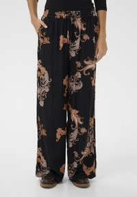 Zwarte wijde broek met bruine en beige paisleypatronen, elastische tailleband met trekkoord, gedragen met bruine schoenen op een effen achtergrond.