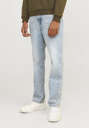 Personne portant un jean droit bleu clair, des baskets blanches et un haut à manches longues vert olive, debout devant un fond clair uni.