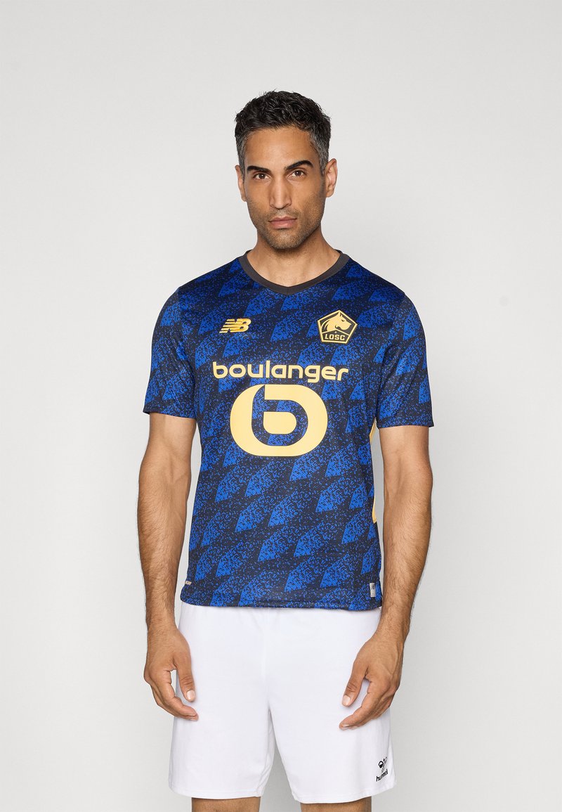 Blauwe voetbalsjerp met driehoekige patronen, voorzien van een gele logo en tekst. Korte mouwen en een ronde halslijn, gecombineerd met witte shorts.
