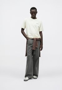 Gaismas dzeltena īsroku t-krekls ar logotipu, pelēkas vaļīgas džinsa bikses ar platiem galiem un brūns džemperis, pārģērbts ap jostasvietu. Balti un pelēki sporta apavi.