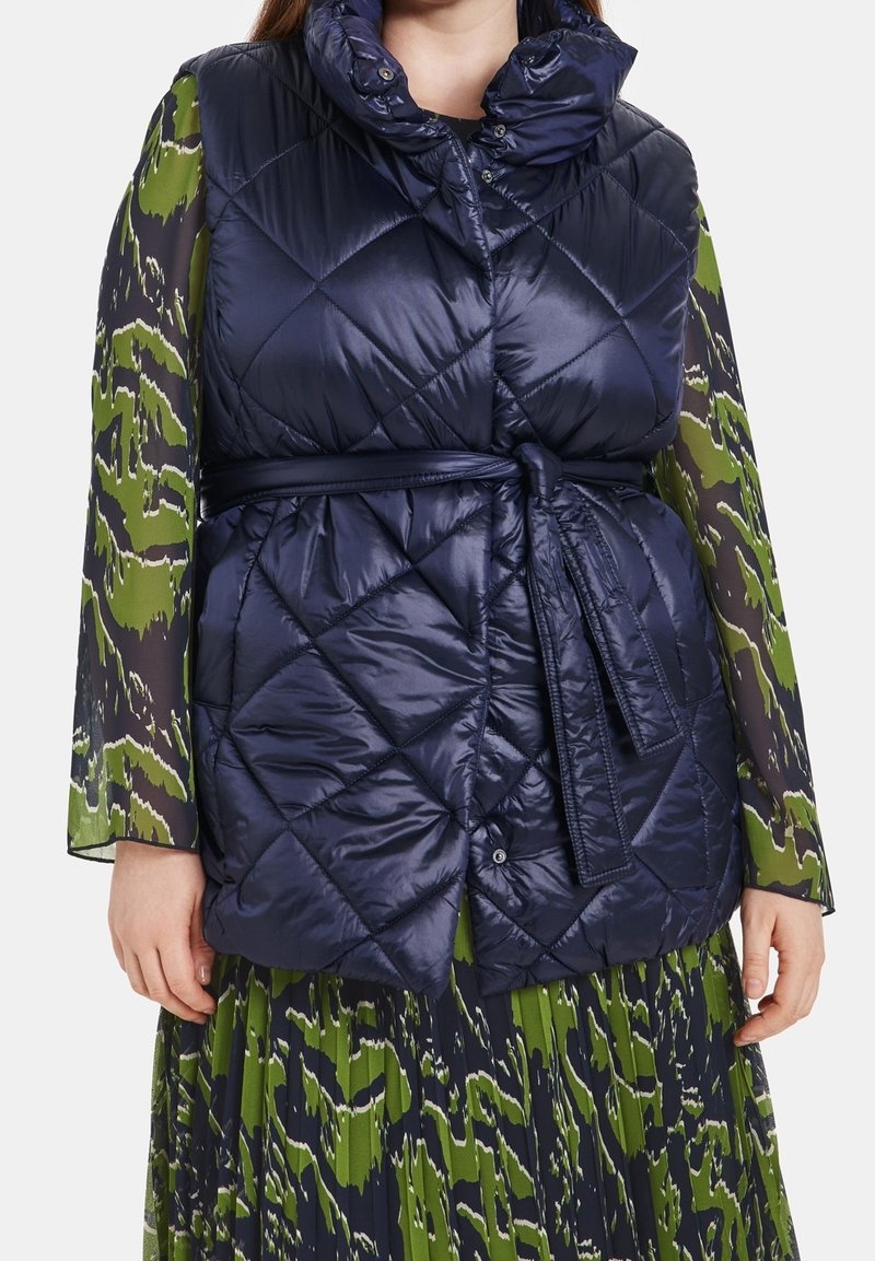 Quilted navy puffer vest with high collar, belted waist, and glossy finish; worn over a long-sleeve green patterned dress.