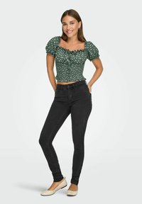 Grøn blomster top med puffærmer og bølgede kanter, parret med sorte tætsiddende jeans og lyse flade sko. Simpelt, afslappet look.