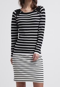 Schwarz-weiß gestreiftes Langarmoberteil mit rundem Ausschnitt, kombiniert mit einem engen schwarz-weiß gestreiften Rock. Auffällige Reißverschlüsse an den Schultern.