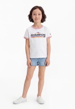 Barn med krøllet hår iført hvid Champion t-shirt med pink kant, denimshorts og hvide sneakers, stående mod en hvid baggrund.