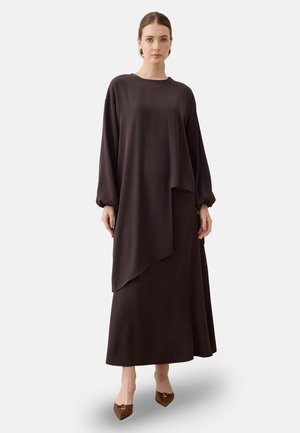 Kvinde iført en lang mørkebrunt asymmetrisk kjole med lange ærmer og brune spidse hæle, stående mod en ensartet lys baggrund.