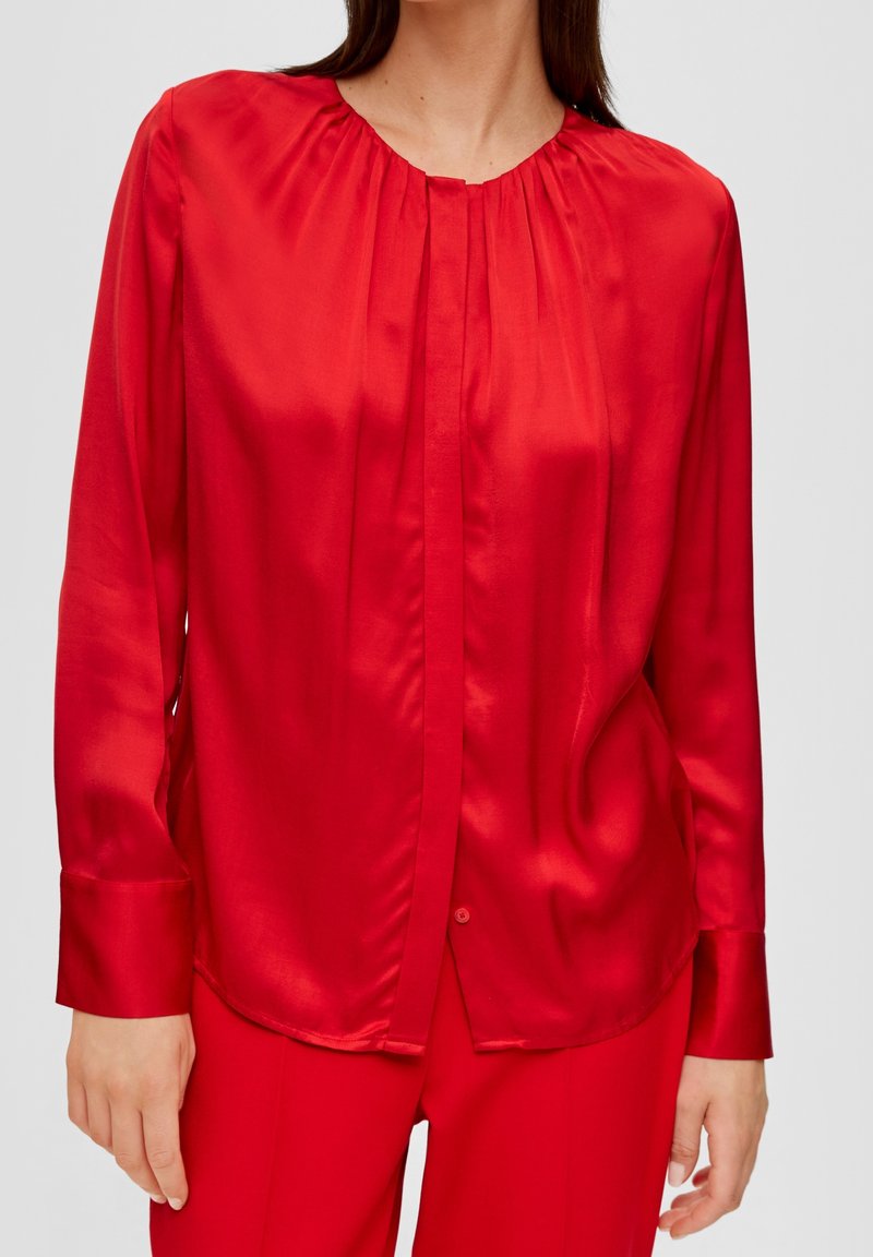 Femme portant un chemisier rouge brillant à manches longues avec des plis près du col et un pantalon rouge assorti, sur un fond uni.