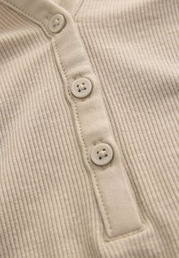 Tejido beige acanalado con tapeta de cinco botones. Cuatro botones planos crema visibles, que muestran una textura suave y un diseño de tejido apretado.