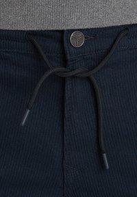 Pantalons en velours côtelé bleu marine avec un cordon de serrage noir et un bouton métallique rond. La texture est côtelée, avec des détails de couture visibles.