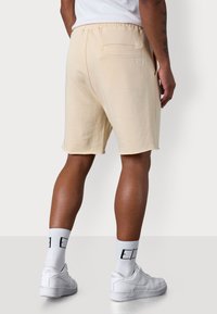 Cream-colored, knee-length shorts with an elastic waistband and rear pocket. Soft fabric with raw edges. Paired with white sneakers and socks.