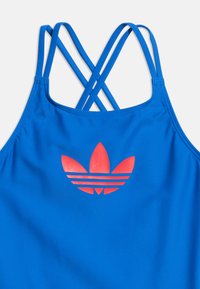 Μπλε αθλητική φανέλα χωρίς μανίκια με λεπτούς διασταυρούμενους ιμάντες και μεγάλο κόκκινο λογότυπο τριφυλλιού Adidas στο στήθος.