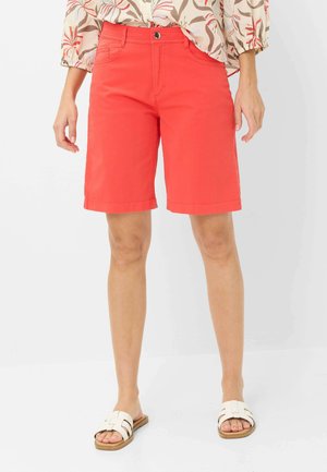 Donna con pantaloncini rossi fino al ginocchio, top floreale e sandali bianchi aperti, in piedi davanti a uno sfondo semplice.