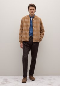 Giacca bomber a quadri marrone con polsini a coste, camicia di jeans blu, pantaloni marrone scuro e scarpe in camoscio marrone. Texture lisce e strutturate.