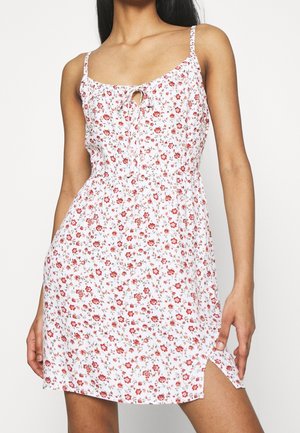 Kvinde iført hvid ærmeløs kjole med rødt blomsterprint og sideslids, stående med den ene hånd, der rører ved kjolens kant.
