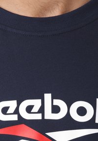 Camiseta de algodón de color azul marino con cuello redondo acanalado y gran logo blanco de "reebok". Presenta detalles rojos y blancos en el diseño.