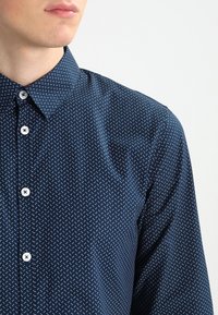 Navy blue button-up shirt with a fine white polka dot pattern, spread collar, and white buttons for closure. Smooth, lightweight fabric texture.