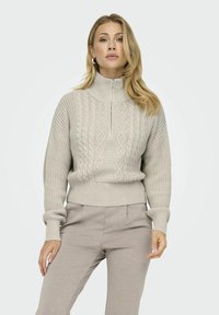 Helles graues Strickpullover mit hohem Kragen, halbem Reißverschluss vorne und strukturiertem Kabelmuster. Gerippte Bündchen und Saum setzen Akzente.