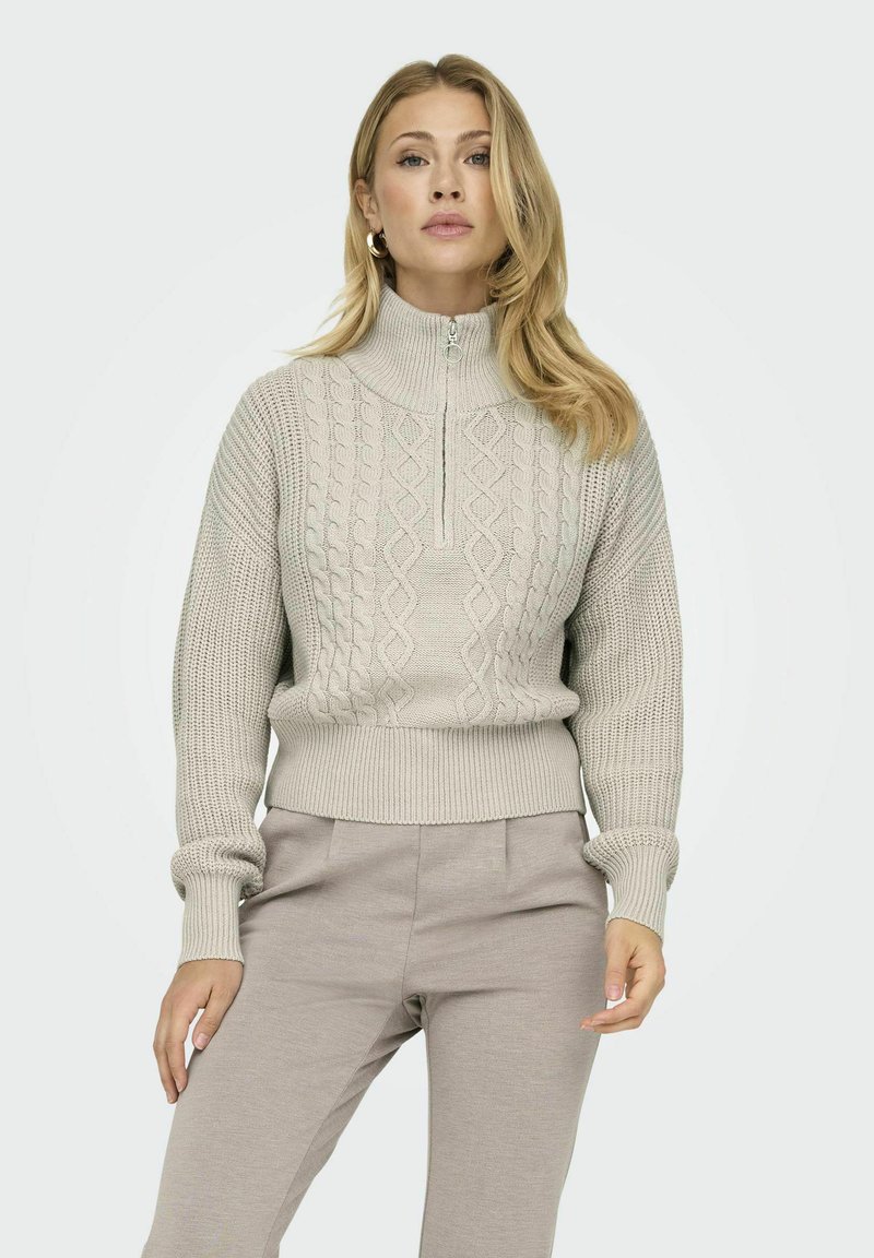 Helles graues Strickpullover mit hohem Kragen, halbem Reißverschluss vorne und strukturiertem Kabelmuster. Gerippte Bündchen und Saum setzen Akzente.