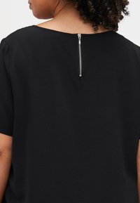 Haut noir à manches courtes avec un tissu texturé et un détail de fermeture éclair argenté visible au dos. Design simple et ample.