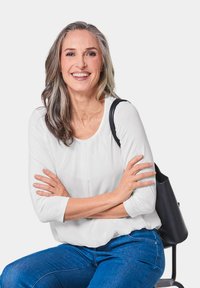 Weiße Bluse mit langen Ärmeln, blaue Jeans und schwarze Schultertasche. Die Person sitzt mit verschränkten Armen und lächelt direkt in die Kamera.