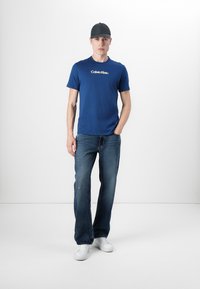 Μπλε μπλουζάκι με στρογγυλή λαιμόκοψη και λευκό λογότυπο "Calvin Klein", συνδυασμένο με χαλαρά, σκούρα τζιν παντελόνια και λευκά αθλητικά παπούτσια, ολοκληρωμένο με ένα μαύρο καπέλο.