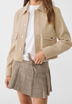 Mujer que lleva una chaqueta de lana beige con botones sobre una camisa blanca, combinada con una falda plisada de cuadros atada con un lazo en la cintura.