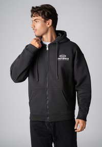 Black zip-up hoodie made of soft fabric, featuring a kangaroo pocket, drawstrings, and printed logo detailing on the chest.