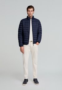 Chaqueta de plumas de color navy con cuello alto, textura acolchada y cierre de cremallera. Lleva puesta una camisa clara, combinada con pantalones crema y zapatillas oscuras.