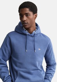 Jonge man met een lichtblauwe capuchonsweater met een voorzak en een klein logo op de borst, poserend tegen een effen witte achtergrond.