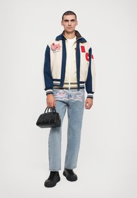Creme og marineblå varsity jakke med mærker, creme t-shirt, lyseblå jeans med pink broderi og en lille sort håndtaske; sorte sneakers.