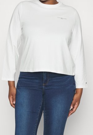 Person trägt ein weißes Langarmshirt von Tommy Hilfiger und blaue Jeans, steht mit entspannt an den Seiten hängenden Händen.