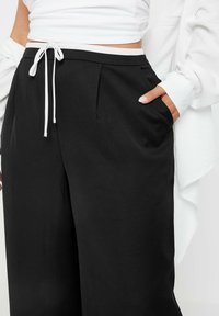 Svarta byxor med hög midja, som har en vit snörning. Tyget har en mjuk textur och veck framtill, med sidofickor synliga.