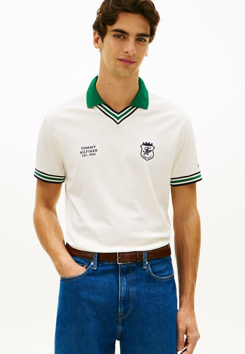 Jeune homme portant un polo blanc avec col vert et poignets rayés, jean bleu et ceinture marron, debout avec une main dans la poche.
