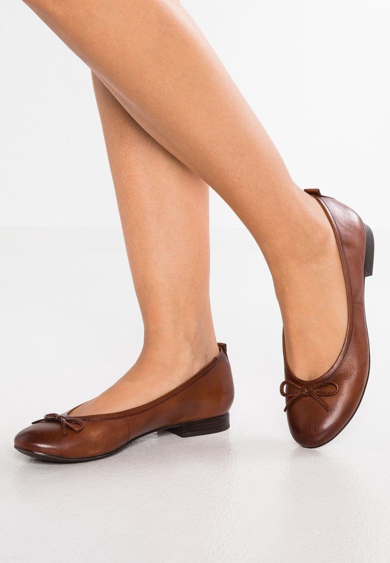 Paire de jambes portant des ballerines en cuir marron avec de petits nœuds, un pied à plat sur le sol, l'autre sur la pointe des pieds.