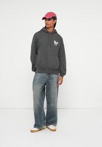 Felpa grigia con grafica di una farfalla, abbinata a jeans a gamba larga azzurri e un cappellino rosso. Sneakers bianche completano il look.