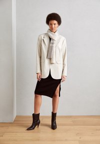 Blazer beige sur mesure porté sur une robe noire côtelée avec une fente latérale. Écharpe rayée grise, bottines noires à bout pointu. Texture de plancher en bois visible.