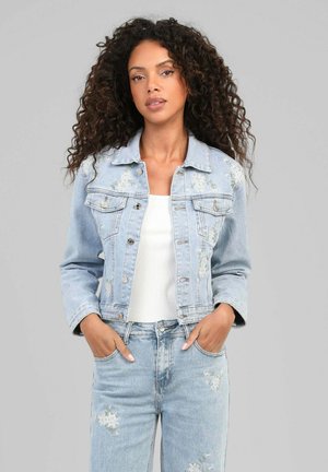 Femme aux cheveux bouclés portant une veste en jean bleu clair brodée de fleurs et un jean assorti sur un haut blanc, mains dans les poches.
