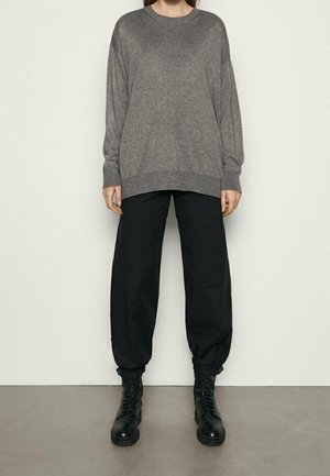 Personne portant un pull gris ample à manches longues, un pantalon noir retroussé et des bottes noires à lacets, debout contre un mur clair uni.