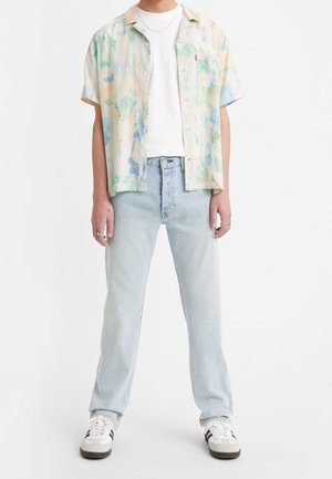 Camisa de botones de manga corta en tonos pastel con un patrón de acuarela, llevada sobre una camiseta blanca y unos jeans de mezclilla azul claro, combinada con zapatillas blancas.